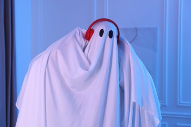 Photo of Funny ghost. Man covered with white sheet listening to music with headphones in blue light at home