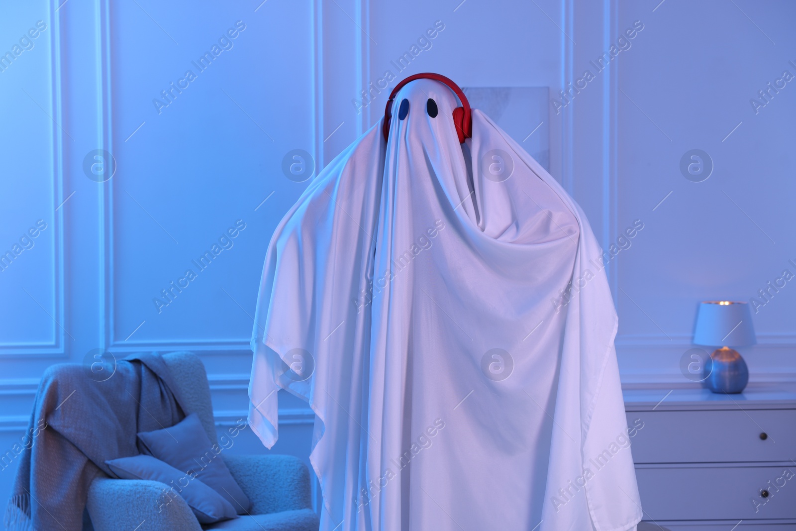 Funny ghost. Man covered with white sheet listening to music with headphones in blue light at home Photo of Funny ghost. Man covered with white sheet listening to music with headphones in blue light at home