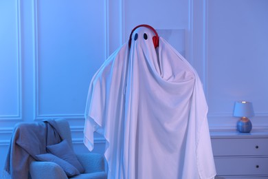 Photo of Funny ghost. Man covered with white sheet listening to music with headphones in blue light at home