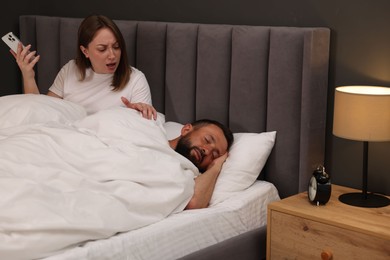 Photo of Betrayal. Jealous woman finding proofs of her unfaithful husband on smartphone while he sleeping and awaken him in bedroom