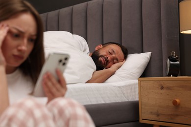 Photo of Betrayal. Frustrated woman finding proofs of her unfaithful husband on smartphone while he sleeping on bed at home, selective focus