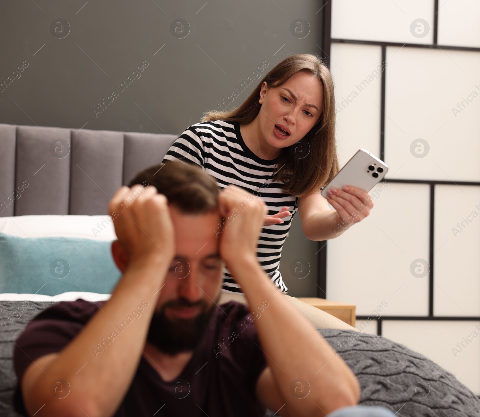 Betrayal. Angry woman with smartphone shouting at her offended husband on bed at home, selective focus Photo of Betrayal. Angry woman with smartphone shouting at her offended husband on bed at home, selective focus