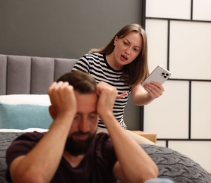 Photo of Betrayal. Angry woman with smartphone shouting at her offended husband on bed at home, selective focus