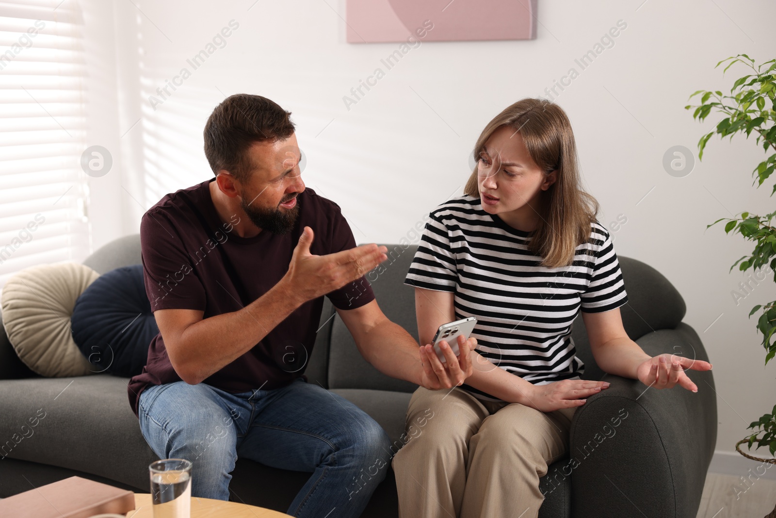 Betrayal. Husband showing smartphone with proofs to his unfaithful wife on sofa at home Photo of Betrayal. Husband showing smartphone with proofs to his unfaithful wife on sofa at home