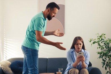 Photo of Betrayal. Offended wife with smartphone ignoring her husband on sofa at home
