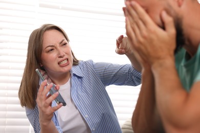 Photo of Betrayal. Emotional wife showing smartphone with proofs to her unfaithful husband at home, selective focus