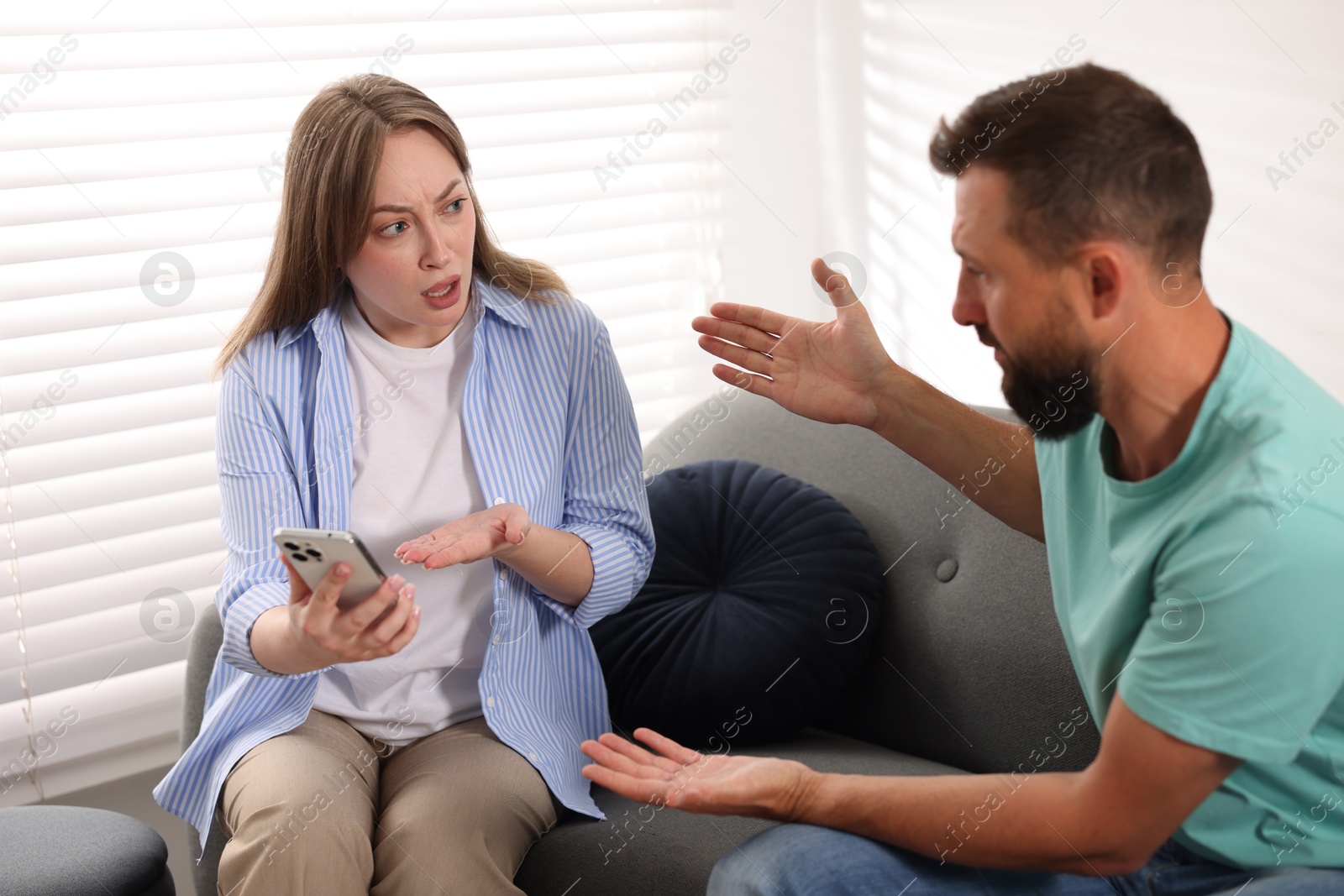 Betrayal. Emotional wife showing smartphone with proofs to her unfaithful husband on sofa at home Photo of Betrayal. Emotional wife showing smartphone with proofs to her unfaithful husband on sofa at home