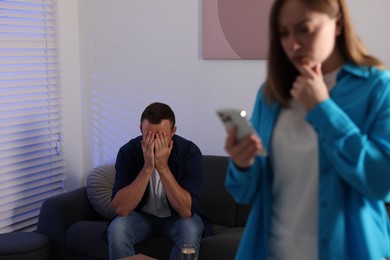Photo of Betrayal.Man covering face with hands while his wife finding proofs of his betrayal on smartphone at home, selective focus