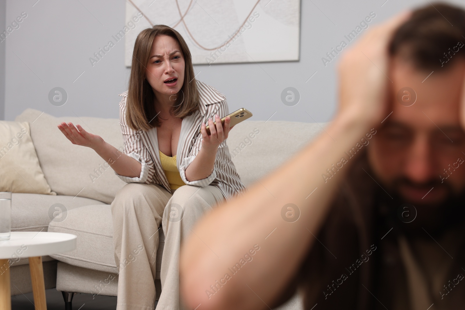 Betrayal. Angry woman with smartphone shouting at her offended husband on sofa at home, selective focus Photo of Betrayal. Angry woman with smartphone shouting at her offended husband on sofa at home, selective focus