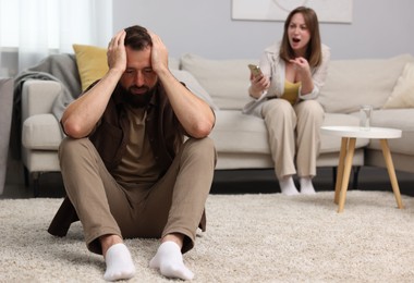 Photo of Betrayal. Angry woman with smartphone shouting at her offended husband on sofa at home, selective focus