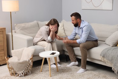 Photo of Betrayal. Husband with glass of water apologizing to his offended wife on sofa at home