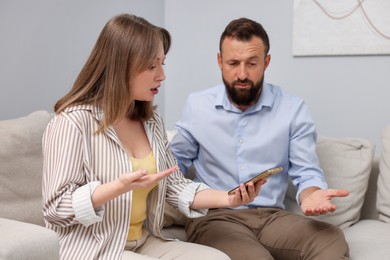 Photo of Betrayal. Wife showing smartphone with proofs to her unfaithful husband on sofa at home
