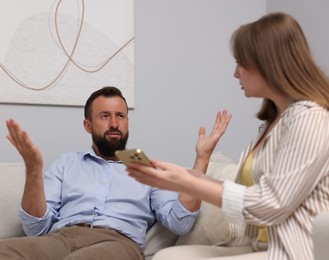 Photo of Betrayal. Wife showing smartphone with proofs to her unfaithful husband on sofa at home