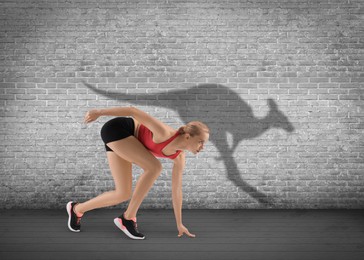 Image of Athletic young woman on starting position for run. Her shadow in form of kangaroo on grey brick wall. Sports poster