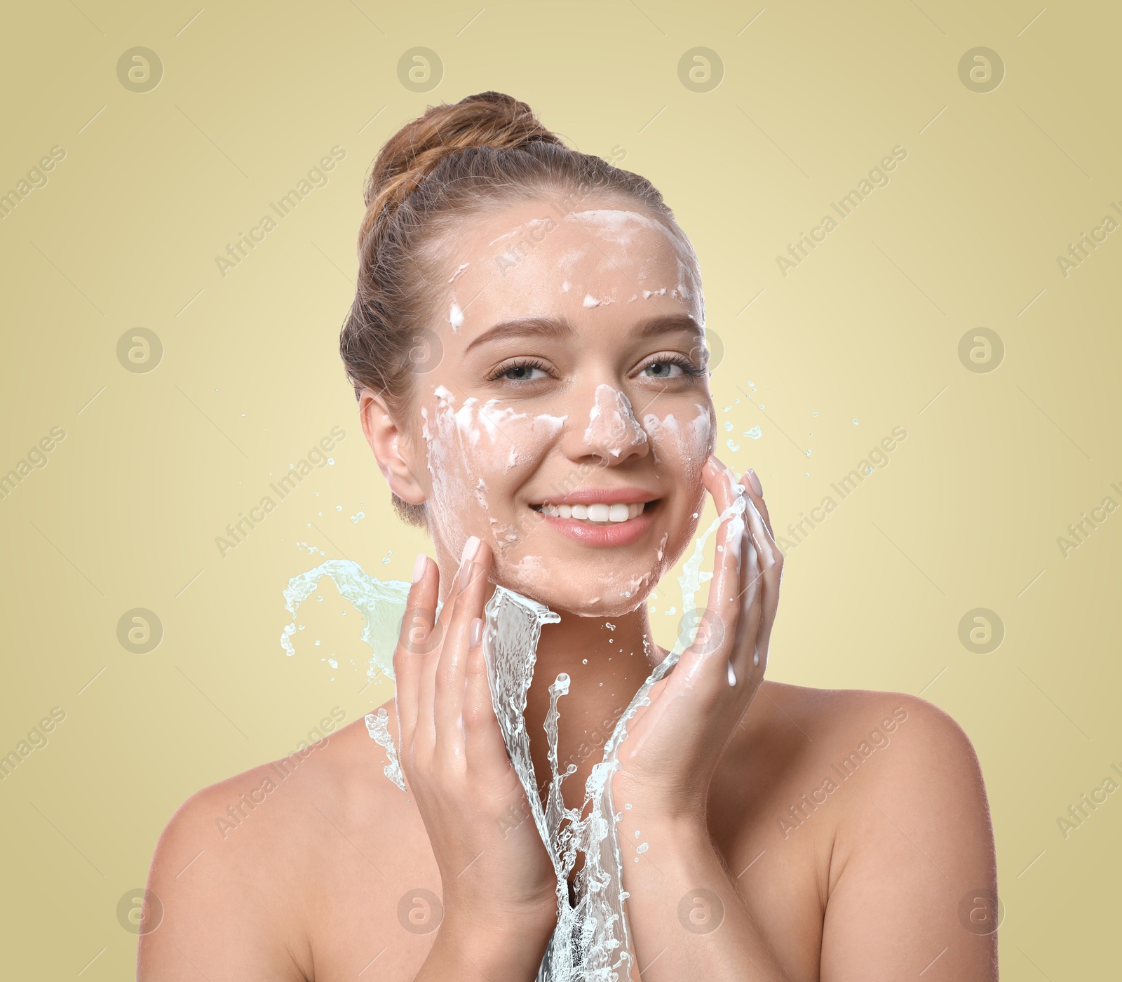 Young woman washing face with cleansing foam and water splash against beige background Image of Young woman washing face with cleansing foam and water splash against beige background
