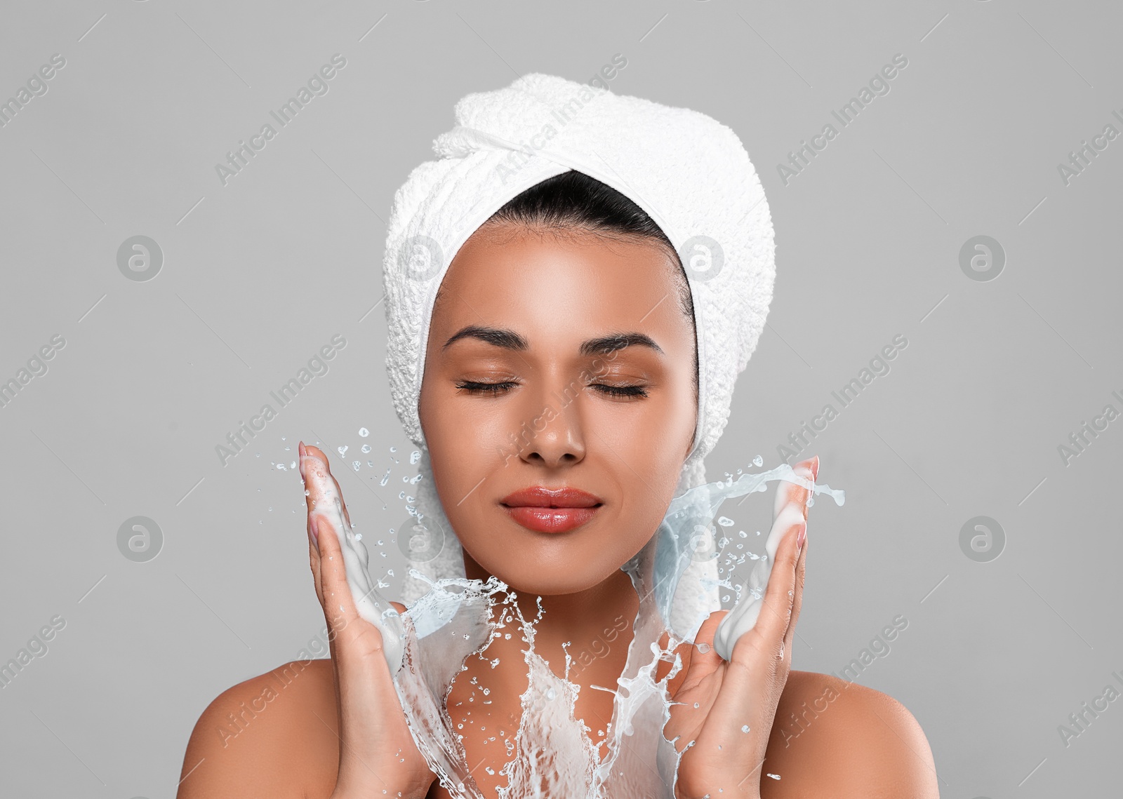 Young woman washing face with cleansing foam and water splash against grey background Image of Young woman washing face with cleansing foam and water splash against grey background