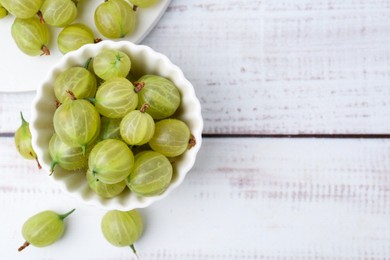 Photo of Fresh green gooseberries in bowl on white wooden table, flat lay. Space for text