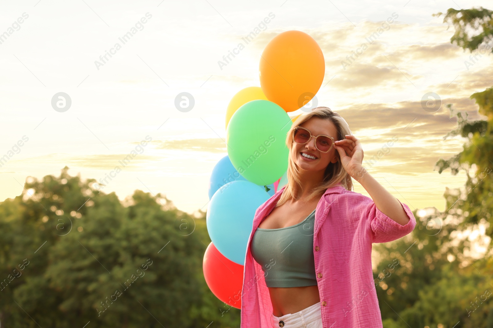 Photo of Smiling woman in sunglasses with bunch of colorful balloons outdoors at sunset. Space for text