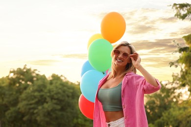 Photo of Smiling woman in sunglasses with bunch of colorful balloons outdoors at sunset. Space for text