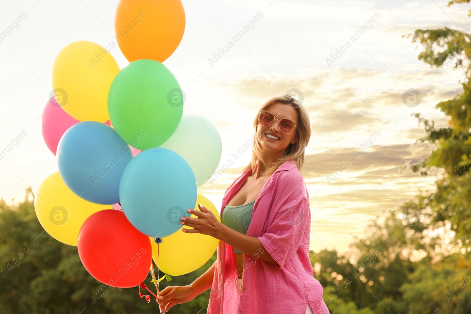 Photo of Smiling woman in sunglasses with bunch of colorful balloons outdoors at sunset