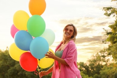 Photo of Smiling woman in sunglasses with bunch of colorful balloons outdoors at sunset