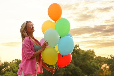 Photo of Smiling woman in sunglasses with bunch of colorful balloons outdoors at sunset