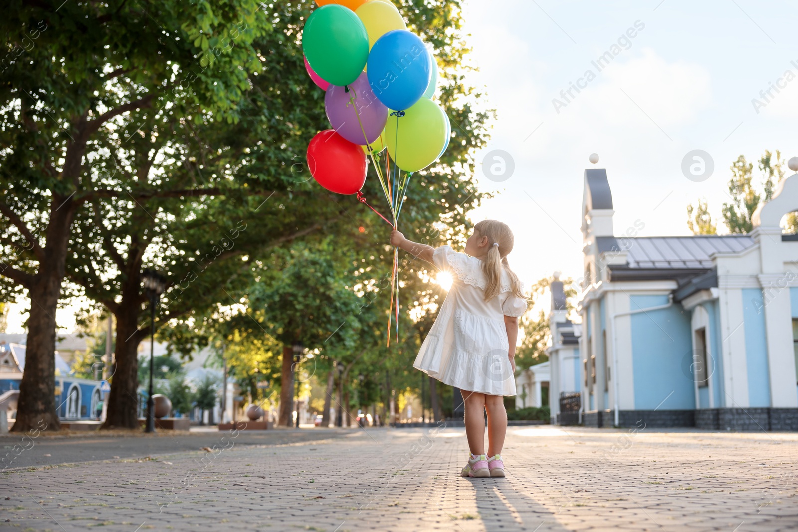 Photo of Little girl in dress with bunch of colorful balloons outdoors at sunset, back view