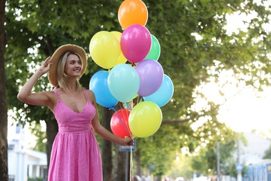 Photo of Smiling woman in hat and dress with bunch of colorful balloons outdoors