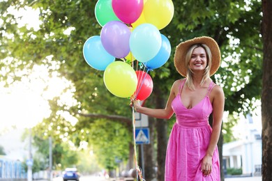 Photo of Portrait of smiling woman in hat and dress with bunch of colorful balloons outdoors