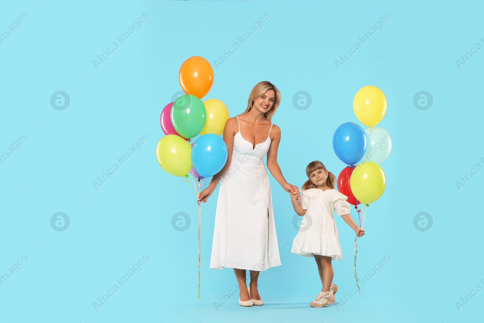 Happy mother and her little daughter with bunches of colorful balloons holding hands on light blue background Photo of Happy mother and her little daughter with bunches of colorful balloons holding hands on light blue background