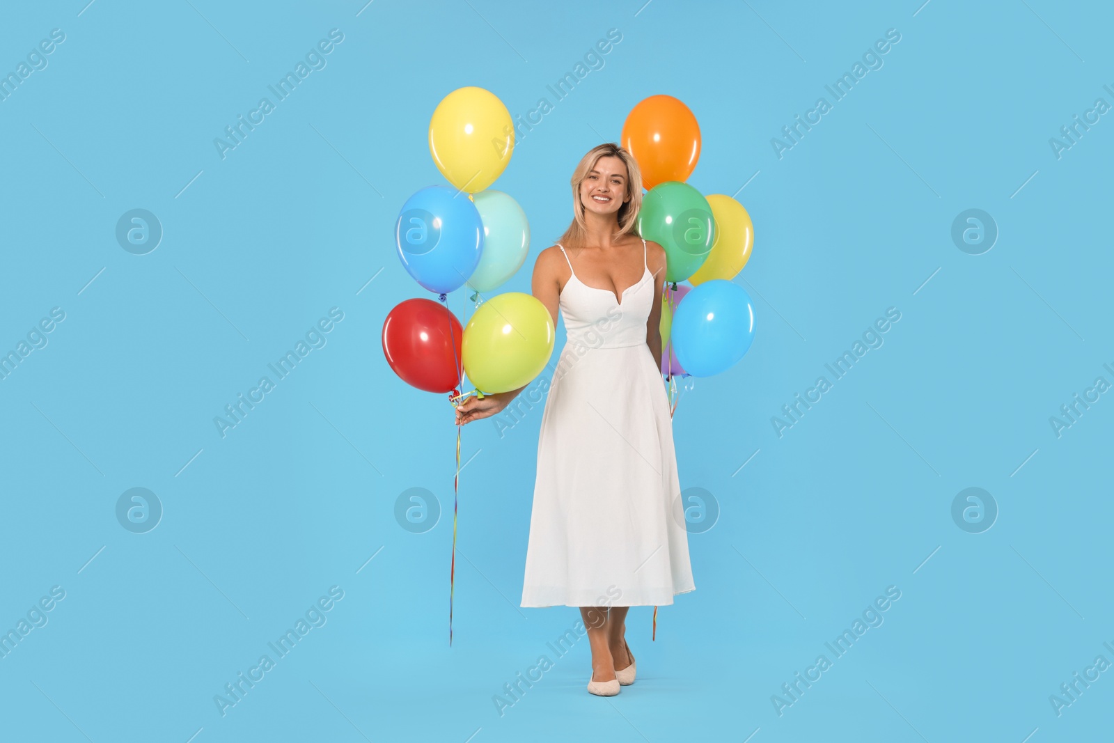 Smiling woman in dress with bunches of colorful balloons on light blue background Photo of Smiling woman in dress with bunches of colorful balloons on light blue background