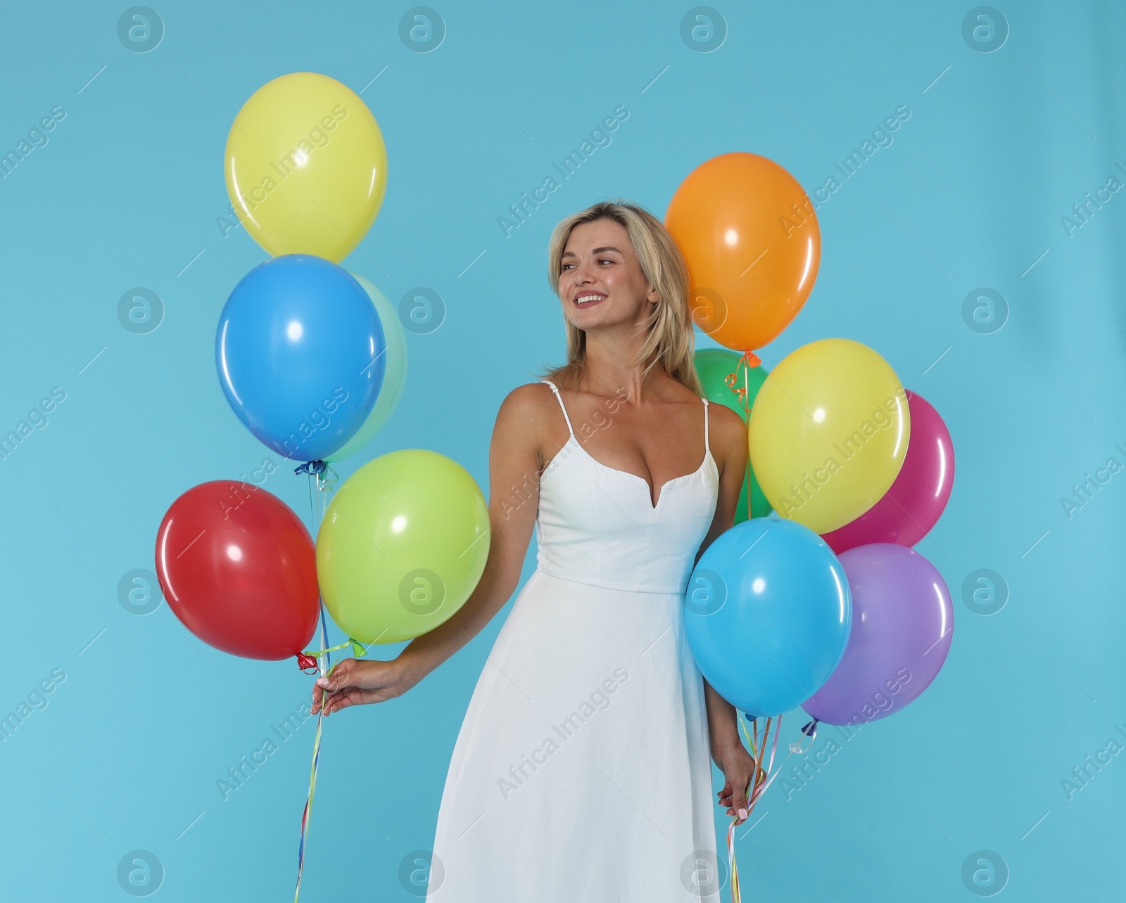 Smiling woman in dress with bunches of colorful balloons on light blue background Photo of Smiling woman in dress with bunches of colorful balloons on light blue background