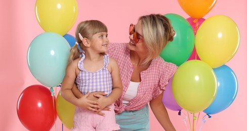 Photo of Happy mother in sunglasses and her little daughter with bunches of colorful balloons on pink background