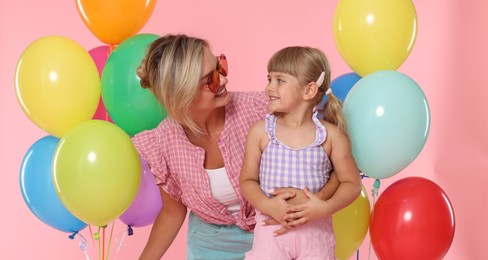 Photo of Happy mother in sunglasses and her little daughter with bunches of colorful balloons on pink background