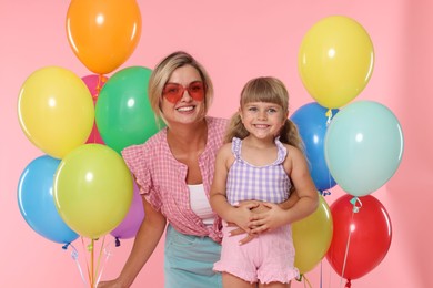 Photo of Happy mother in sunglasses and her little daughter with bunches of colorful balloons on pink background