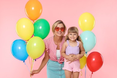 Photo of Happy mother in sunglasses and her little daughter with bunches of colorful balloons on pink background