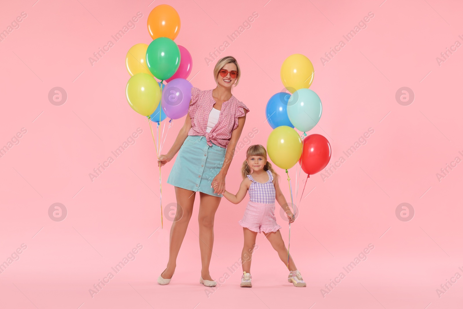 Happy mother in sunglasses and her little daughter with bunches of colorful balloons holding hands on pink background Photo of Happy mother in sunglasses and her little daughter with bunches of colorful balloons holding hands on pink background