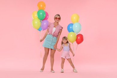 Photo of Happy mother in sunglasses and her little daughter with bunches of colorful balloons holding hands on pink background