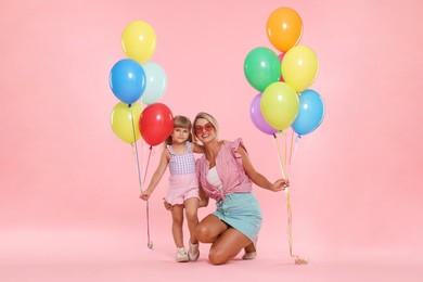 Photo of Happy mother in sunglasses and her little daughter with bunches of colorful balloons on pink background