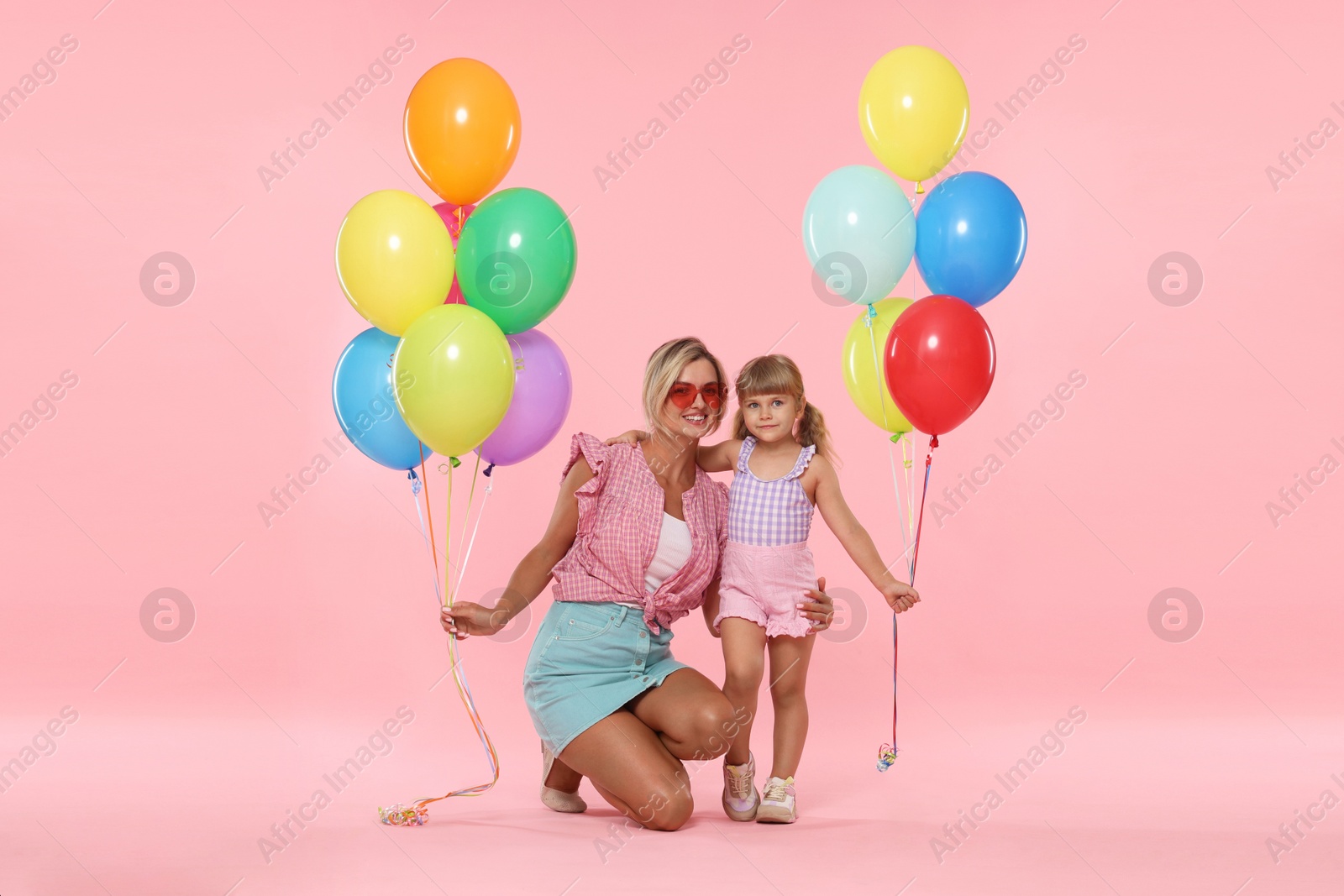 Photo of Happy mother in sunglasses and her little daughter with bunches of colorful balloons on pink background