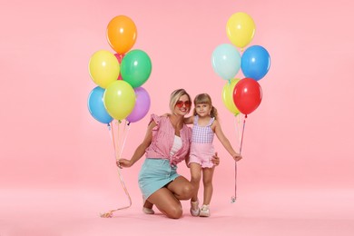 Photo of Happy mother in sunglasses and her little daughter with bunches of colorful balloons on pink background