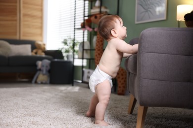 Photo of First steps. Cute little baby in diaper near sofa at home, space for text