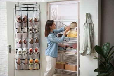 Photo of Woman taking cap from shelf near organizer with different footwear at home. Shoe storage