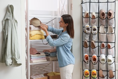 Photo of Woman taking cap from shelf near organizer with different footwear at home. Shoe storage