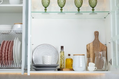 Photo of Different dishware, glasses and other kitchen utensils on shelves in cabinets indoors