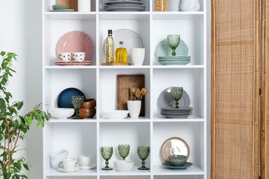 Photo of Shelving unit with different dishware, glasses and other kitchen utensils near white wall indoors