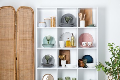 Photo of Shelving unit with different dishware, glasses and other kitchen utensils near white wall indoors