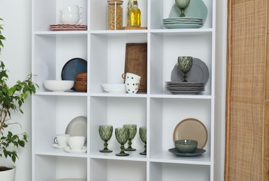 Photo of Shelving unit with different dishware, glasses and other kitchen utensils near white wall indoors