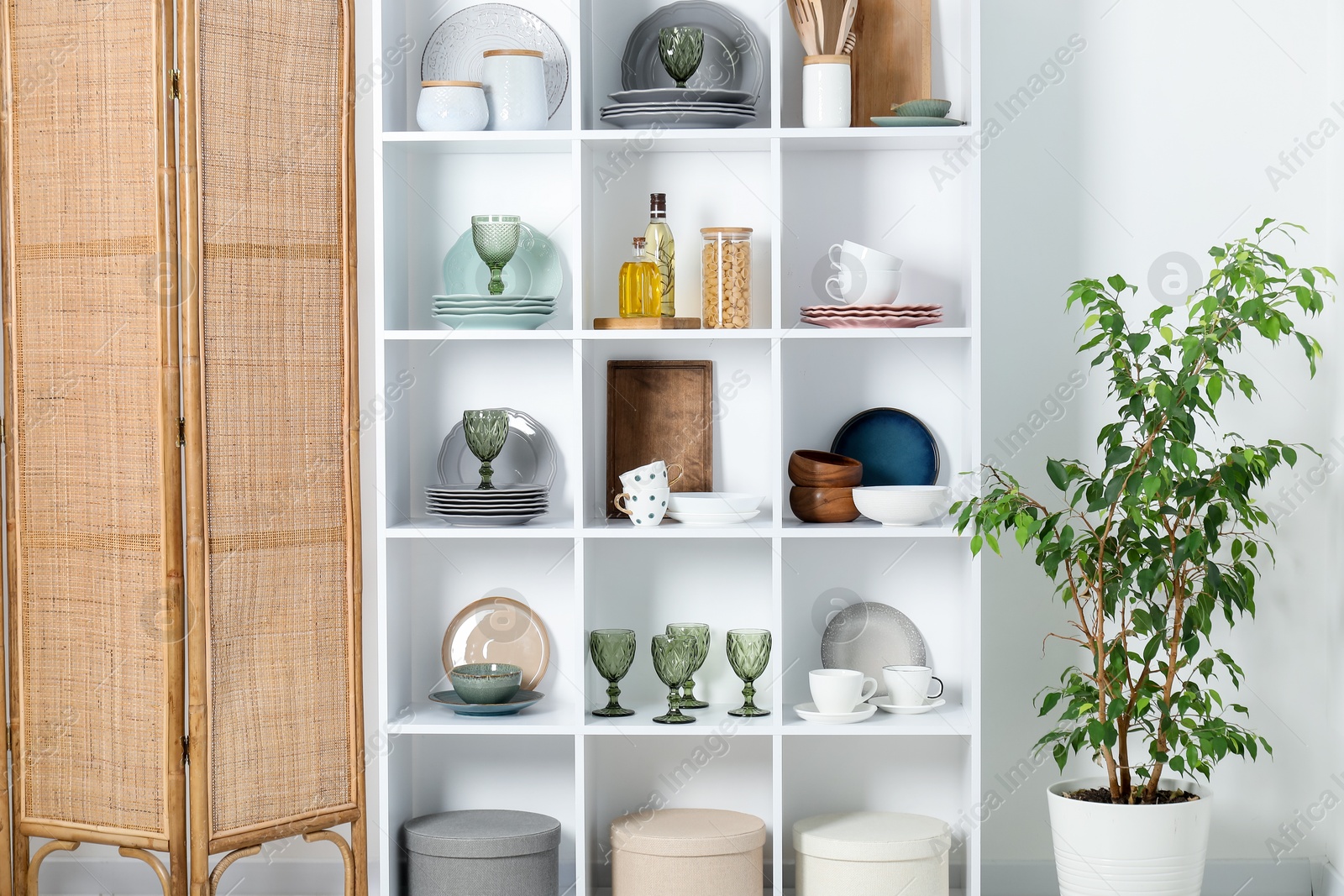 Photo of Shelving unit with different dishware, glasses and other kitchen utensils near white wall indoors