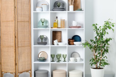 Photo of Shelving unit with different dishware, glasses and other kitchen utensils near white wall indoors